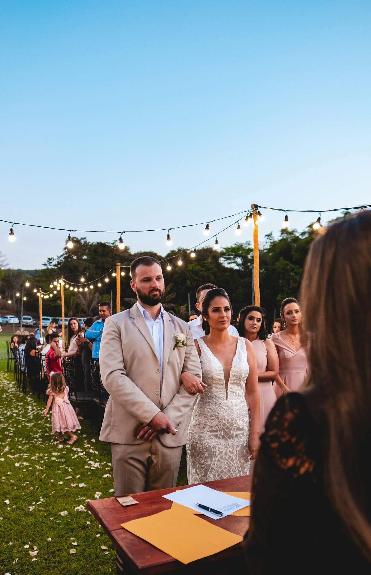 A celebrant leading an outdoor ceremony at dusk, the couple and their guests beyond the signing table
